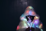 A person wearing a fluffy, white fur coat with a large hood that casts colorful, ambient lighting across the fabric. The scene is set against a dark background, creating a contrast that highlights the soft texture of the fur and the glow of the lights.