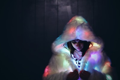 A person wearing a fluffy, white fur coat with a large hood that casts colorful, ambient lighting across the fabric. The scene is set against a dark background, creating a contrast that highlights the soft texture of the fur and the glow of the lights.