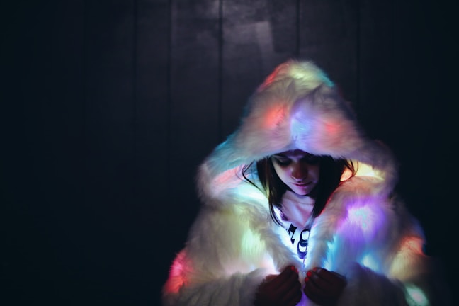 A person wearing a fluffy, white fur coat with a large hood that casts colorful, ambient lighting across the fabric. The scene is set against a dark background, creating a contrast that highlights the soft texture of the fur and the glow of the lights.