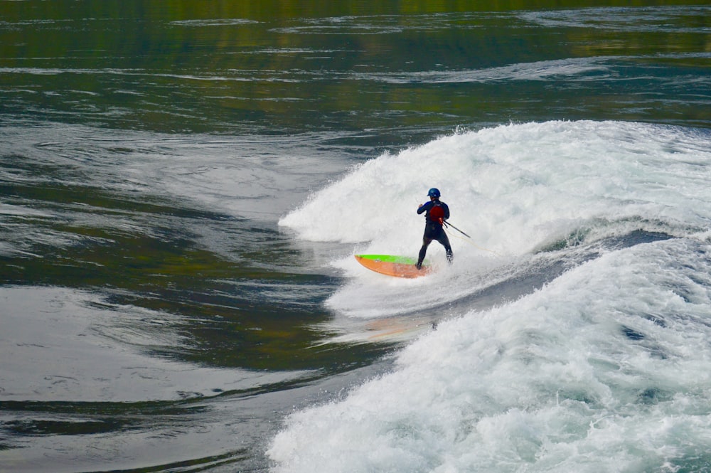 Persona de pie sobre una tabla de surf naranja y verde en la ola del mar