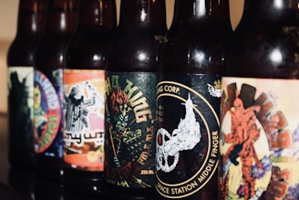 Several beer bottles stand in a row, each featuring unique and colorful labels with various artistic designs and imagery. The labels include elements like abstract patterns, illustrations, and text with different fonts.