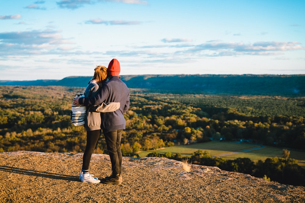 man and woman standing on mountain cliff in front of green trees