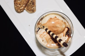 A glass dish filled with vanilla ice cream, drizzled with caramel sauce and garnished with a striped chocolate wafer stick. The dish is placed on a white rectangular plate, which also holds two almond biscotti in the top left corner. The background appears to be solid black, providing a contrast to the white plate and the dessert.