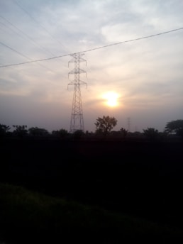A tall electricity transmission tower stands silhouetted against the backdrop of a sunset sky, surrounded by scattered clouds. The sun is partially visible near the horizon, casting a warm glow. Trees and vegetation form a dark silhouette in the foreground.