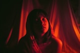 A person with closed eyes is illuminated by a warm, reddish light that casts dramatic shadows around them. The background is soft and draped, adding to the intimate and serene atmosphere.