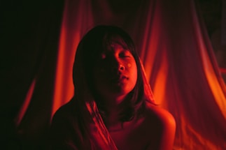 A person with closed eyes is illuminated by a warm, reddish light that casts dramatic shadows around them. The background is soft and draped, adding to the intimate and serene atmosphere.