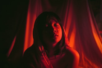 A person with closed eyes is illuminated by a warm, reddish light that casts dramatic shadows around them. The background is soft and draped, adding to the intimate and serene atmosphere.