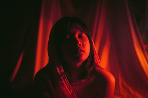 A person with closed eyes is illuminated by a warm, reddish light that casts dramatic shadows around them. The background is soft and draped, adding to the intimate and serene atmosphere.