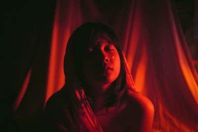 A person with closed eyes is illuminated by a warm, reddish light that casts dramatic shadows around them. The background is soft and draped, adding to the intimate and serene atmosphere.