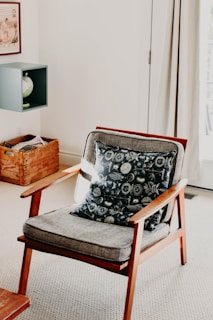 A mid-century style wooden armchair with a gray cushion and a patterned throw pillow sits on a light carpeted floor. To the left, a wooden crate partially filled with newspapers rests on the ground. Above the crate, a small wall-mounted shelf holds a decorative globe. The room has a neutral and bright aesthetic with a framed picture on the wall and curtains partially covering a window or door.