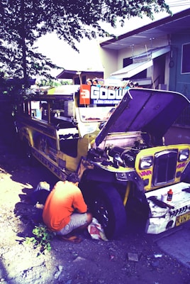A person in an orange shirt is crouched down, working on the wheel of a yellow jeepney parked by the side of the road. The jeepney's hood is open, and various tools and containers are scattered around. It's situated near a light-colored building and some vegetation.