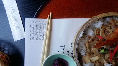 A Japanese meal is presented on a table with a bowl of rice topped with cooked fish and shrimp. A pair of chopsticks rests on a sheet of paper containing Japanese text. A small dish holds pickled red cabbage, and a chopstick wrapper with illustrations and the words 'bamboo chopstick' is visible.