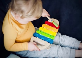 A child wearing a yellow long-sleeve shirt and gray pants is playing with a colorful xylophone. The xylophone features rainbow-colored bars with small symbols on each. The child is using a mallet to strike the bars.
