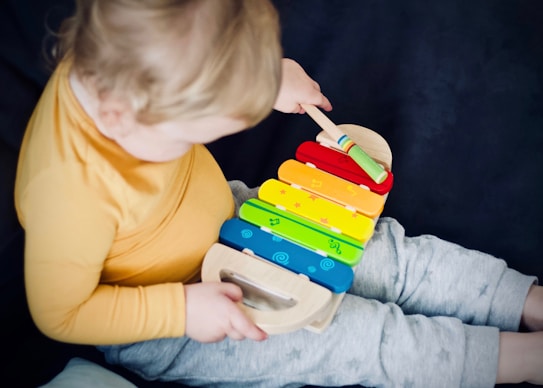 A child wearing a yellow long-sleeve shirt and gray pants is playing with a colorful xylophone. The xylophone features rainbow-colored bars with small symbols on each. The child is using a mallet to strike the bars.