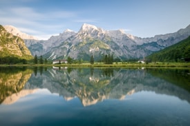 A serene lake mirrors a striking mountain range under a clear blue sky. Pine trees and green hills frame the lake, while a few quaint buildings are nestled amidst the lush landscape. The calm water creates a perfect reflection of the surrounding scenery, enhancing the tranquility of the setting.