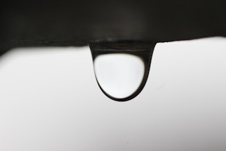 A solitary water droplet hangs precariously from the edge of a dark surface, with a softly blurred background. The droplet is clear, refracting and reflecting the light in a subtle manner.