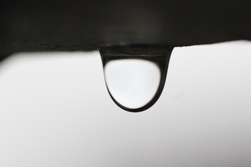A solitary water droplet hangs precariously from the edge of a dark surface, with a softly blurred background. The droplet is clear, refracting and reflecting the light in a subtle manner.