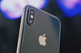 A close-up of a smartphone displaying the Apple logo prominently. The dual camera lenses and flash are clearly visible on the upper left corner of the sleek metallic body. The background is blurred, featuring soft hues of blues and purples, creating a bokeh effect.