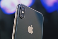 A close-up of a smartphone displaying the Apple logo prominently. The dual camera lenses and flash are clearly visible on the upper left corner of the sleek metallic body. The background is blurred, featuring soft hues of blues and purples, creating a bokeh effect.
