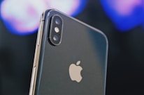 A close-up of a smartphone displaying the Apple logo prominently. The dual camera lenses and flash are clearly visible on the upper left corner of the sleek metallic body. The background is blurred, featuring soft hues of blues and purples, creating a bokeh effect.