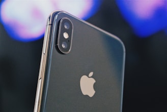 A close-up of a smartphone displaying the Apple logo prominently. The dual camera lenses and flash are clearly visible on the upper left corner of the sleek metallic body. The background is blurred, featuring soft hues of blues and purples, creating a bokeh effect.