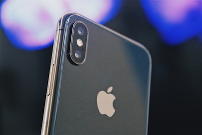 A close-up of a smartphone displaying the Apple logo prominently. The dual camera lenses and flash are clearly visible on the upper left corner of the sleek metallic body. The background is blurred, featuring soft hues of blues and purples, creating a bokeh effect.