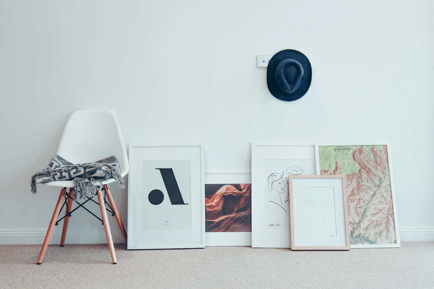 Five framed prints of different sizes leaning against a white wall beside a white chair, waiting to be hung