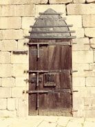 A sturdy, rustic wooden door reinforced with metal strips and bolts, set within an old stone wall. The door features an arched metal design at the top and has multiple locks and latches for security.