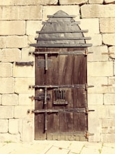 A sturdy, rustic wooden door reinforced with metal strips and bolts, set within an old stone wall. The door features an arched metal design at the top and has multiple locks and latches for security.