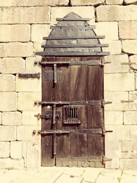 A sturdy, rustic wooden door reinforced with metal strips and bolts, set within an old stone wall. The door features an arched metal design at the top and has multiple locks and latches for security.