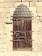 A sturdy, rustic wooden door reinforced with metal strips and bolts, set within an old stone wall. The door features an arched metal design at the top and has multiple locks and latches for security.