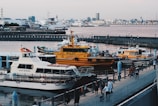 A waterfront scene featuring multiple boats docked at a pier. The most prominent boat is a yellow pilot boat, with the word 'PILOT' written on it. Surrounding it are other smaller boats and yachts. In the background, there are various buildings and structures including larger ships. Several people can be seen walking along the promenade.