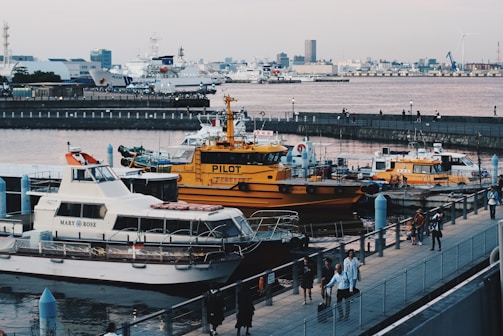 A waterfront scene featuring multiple boats docked at a pier. The most prominent boat is a yellow pilot boat, with the word 'PILOT' written on it. Surrounding it are other smaller boats and yachts. In the background, there are various buildings and structures including larger ships. Several people can be seen walking along the promenade.