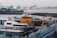 A waterfront scene featuring multiple boats docked at a pier. The most prominent boat is a yellow pilot boat, with the word 'PILOT' written on it. Surrounding it are other smaller boats and yachts. In the background, there are various buildings and structures including larger ships. Several people can be seen walking along the promenade.