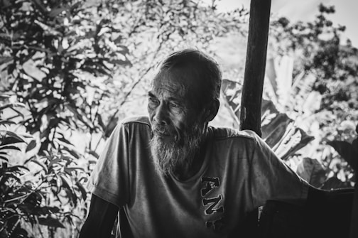 A black and white portrait of an elderly man with a long beard, sitting outdoors. He appears to be wearing a t-shirt and is positioned in front of lush vegetation, including trees and large leaves. The lighting creates deep shadows and highlights his facial features.