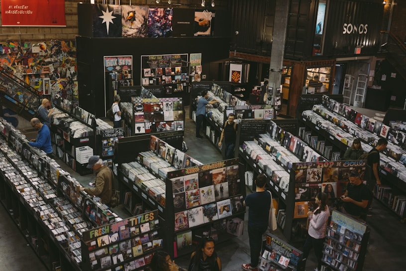A vintage-style record store filled with rows of vinyl records and CDs. People are browsing through the music collections, with walls adorned with posters and album art. The setting appears spacious, with a cozy and nostalgic ambiance.