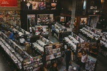 A vintage-style record store filled with rows of vinyl records and CDs. People are browsing through the music collections, with walls adorned with posters and album art. The setting appears spacious, with a cozy and nostalgic ambiance.