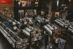 A vintage-style record store filled with rows of vinyl records and CDs. People are browsing through the music collections, with walls adorned with posters and album art. The setting appears spacious, with a cozy and nostalgic ambiance.