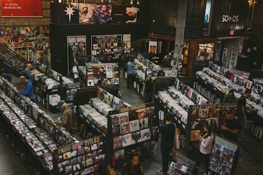 A vintage-style record store filled with rows of vinyl records and CDs. People are browsing through the music collections, with walls adorned with posters and album art. The setting appears spacious, with a cozy and nostalgic ambiance.