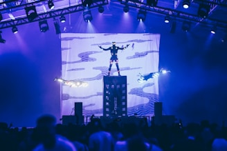 Three individuals are performing an aerial stunt with bright lights, suspended above a crowd in a large indoor venue with structured lighting overhead. A backdrop with abstract patterns is visible, providing a dramatic visual element to the performance.