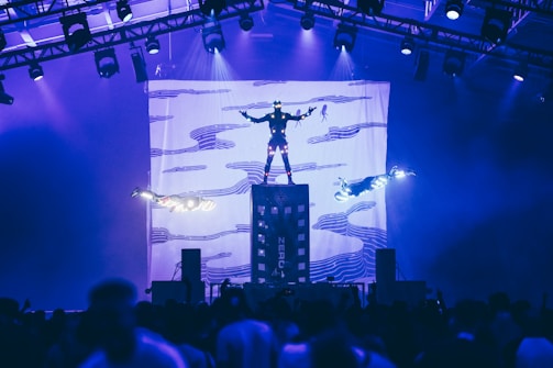 Three individuals are performing an aerial stunt with bright lights, suspended above a crowd in a large indoor venue with structured lighting overhead. A backdrop with abstract patterns is visible, providing a dramatic visual element to the performance.