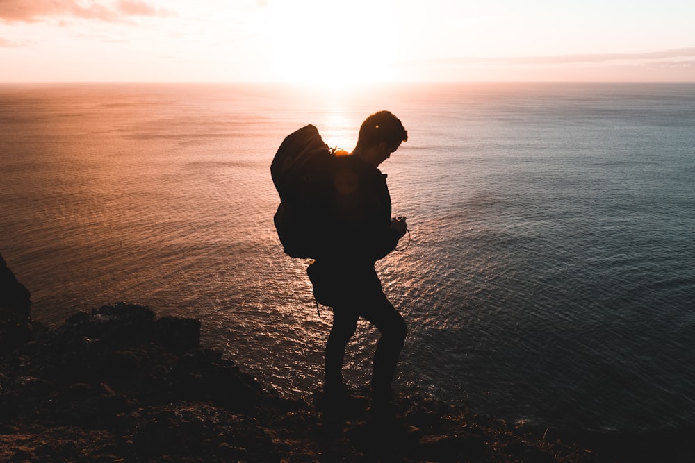 silhouette photo of man standing on rock near body of water