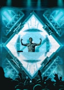 A DJ stands behind a mixing console in front of a large, illuminated LED screen displaying abstract geometric patterns. The DJ is making hand gestures with both hands raised. The crowd in front is energetic, with some people raising their hands. The scene is vibrant and dynamic, with lighting rigs visible on the sides.