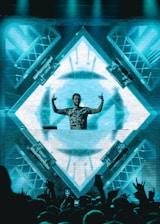 A DJ stands behind a mixing console in front of a large, illuminated LED screen displaying abstract geometric patterns. The DJ is making hand gestures with both hands raised. The crowd in front is energetic, with some people raising their hands. The scene is vibrant and dynamic, with lighting rigs visible on the sides.