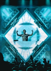 A DJ stands behind a mixing console in front of a large, illuminated LED screen displaying abstract geometric patterns. The DJ is making hand gestures with both hands raised. The crowd in front is energetic, with some people raising their hands. The scene is vibrant and dynamic, with lighting rigs visible on the sides.