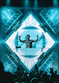 A DJ stands behind a mixing console in front of a large, illuminated LED screen displaying abstract geometric patterns. The DJ is making hand gestures with both hands raised. The crowd in front is energetic, with some people raising their hands. The scene is vibrant and dynamic, with lighting rigs visible on the sides.