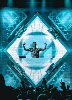 A DJ stands behind a mixing console in front of a large, illuminated LED screen displaying abstract geometric patterns. The DJ is making hand gestures with both hands raised. The crowd in front is energetic, with some people raising their hands. The scene is vibrant and dynamic, with lighting rigs visible on the sides.
