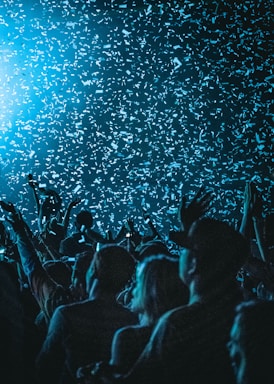 A large crowd of people is gathered, with their hands raised, under a shower of confetti. The atmosphere is vibrant and celebratory, with blue lighting creating a dynamic mood and emphasizing the falling confetti.