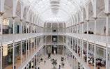 A spacious interior of a large, open museum hall featuring an arched, glass ceiling that allows natural light to fill the area. The architecture is detailed with white pillars and ornate railings. Visitors can be seen walking and observing various exhibits, with some people sitting at a cafe area on the right side.