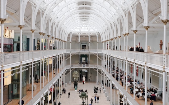 A spacious interior of a large, open museum hall featuring an arched, glass ceiling that allows natural light to fill the area. The architecture is detailed with white pillars and ornate railings. Visitors can be seen walking and observing various exhibits, with some people sitting at a cafe area on the right side.